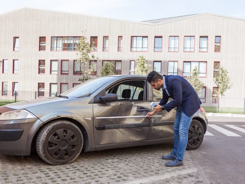 Profile of brunette man wearing jeans and jacket, car inspection for insurance claim the automobile, assesses the damage to the vehicle, examines carefully bent auto door. Outdoor shot.